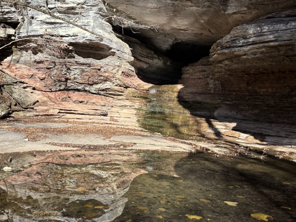 Shallow pool of clear water reflecting layered rock cliffs and the entrance to a dark cave along Lost Valley Trail.