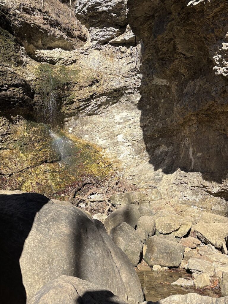 Small waterfall trickling down a rocky cliff into a mostly dry creek bed filled with large boulders and scattered water pools.